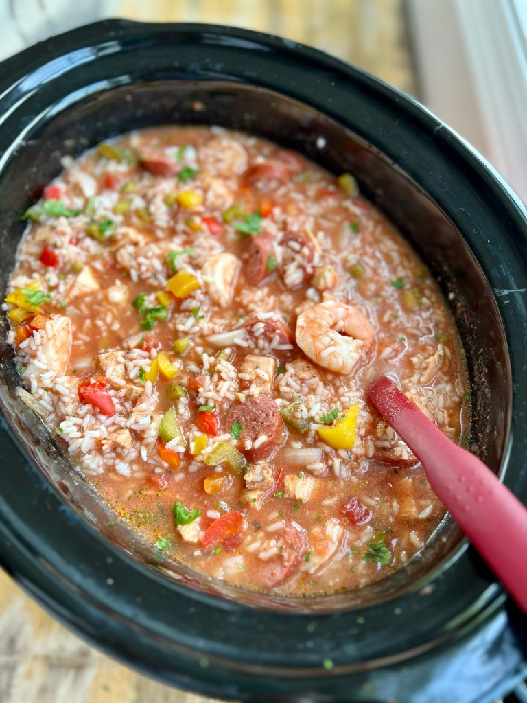 A crockpot filled with jambalaya on a wooden table with a red spoon in it