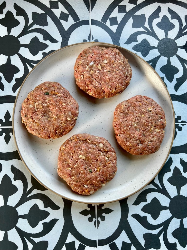 Four raw burger patties on a white plate on a wooden countertop