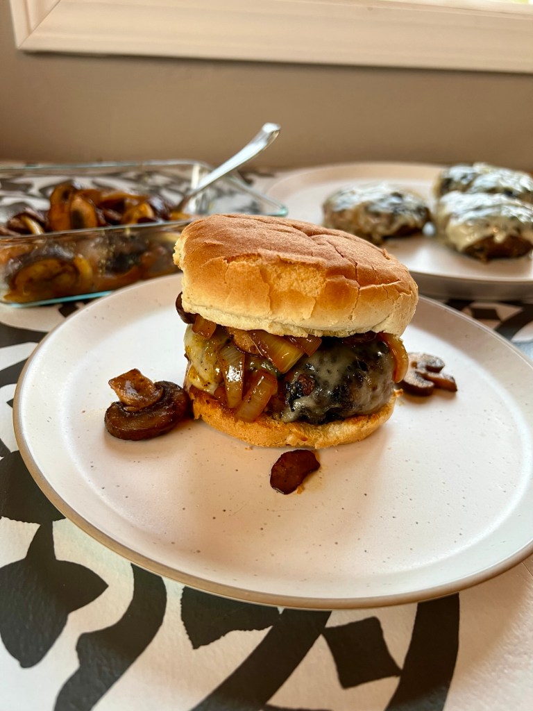 A mushroom and swiss burger on a white plate with a plate of more burgers behind and a dish of mushrooms nearby