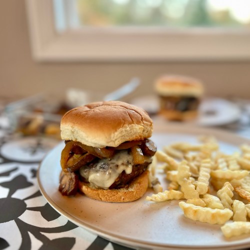A cheeseburger with mushrooms and onions on it with some fries next to it on a white plate with some more burgers and mushrooms in a dish nearby