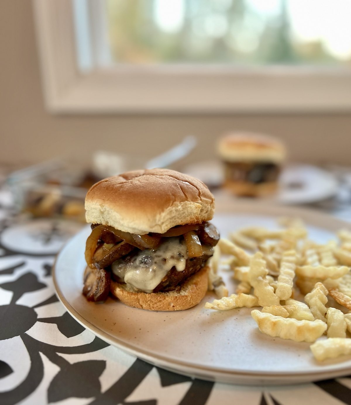 A cheeseburger with mushrooms and onions on it with some fries next to it on a white plate with some more burgers and mushrooms in a dish nearby