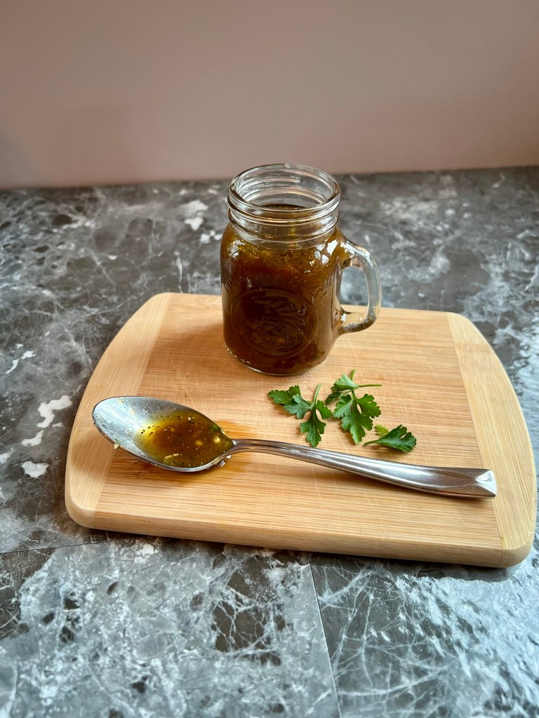 A vinaigrette in a small jar with a spoonful of it and some parsley next to it on a wooden cutting board