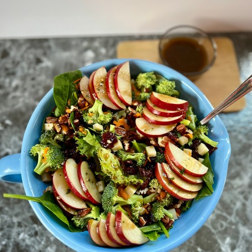 Apple salad in a large blue bowl with a spoon inside and some vinaigrette behind it
