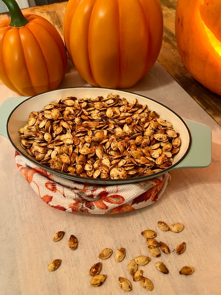 Lemon Pepper Pumpkin Seeds in a glass dish with some pumpkins behind and some seeds scattered around