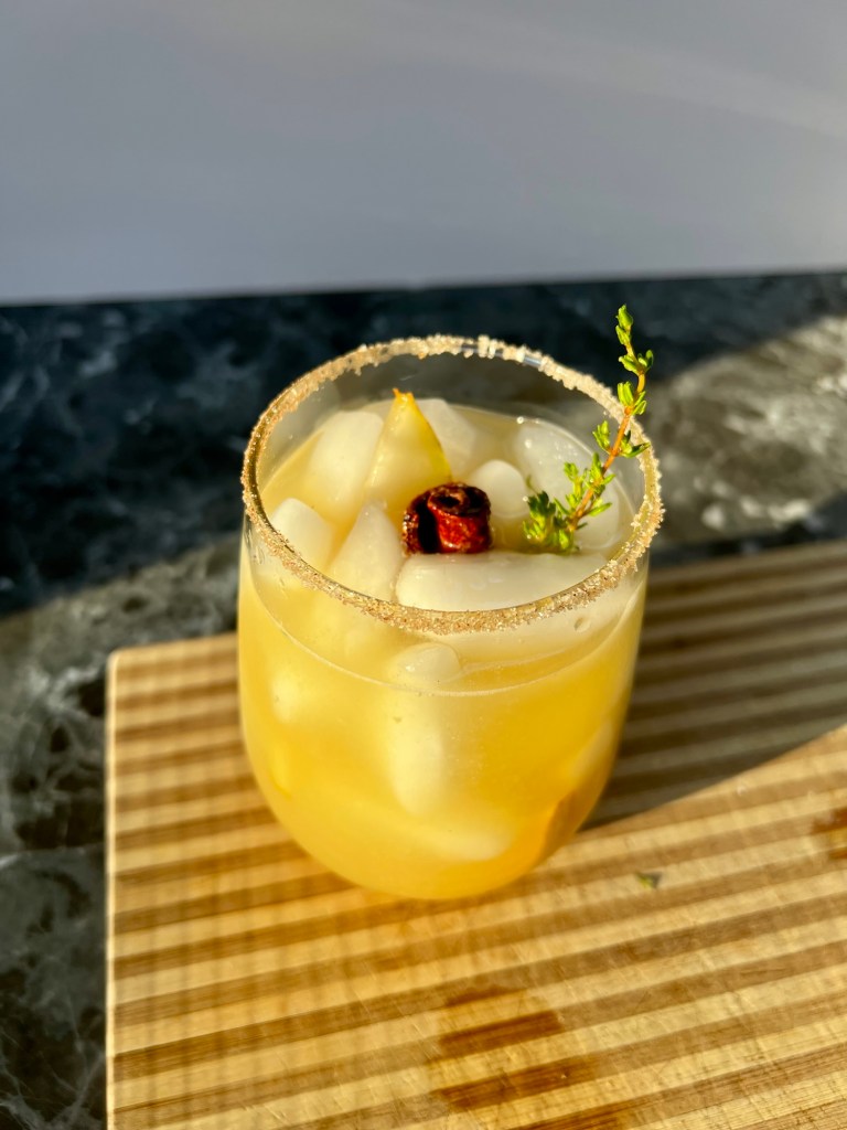 Pear bourbon smash drink with thyme, cinnamon and sugar, a cinnamon stick and some pear for garnish on a wooden cutting board