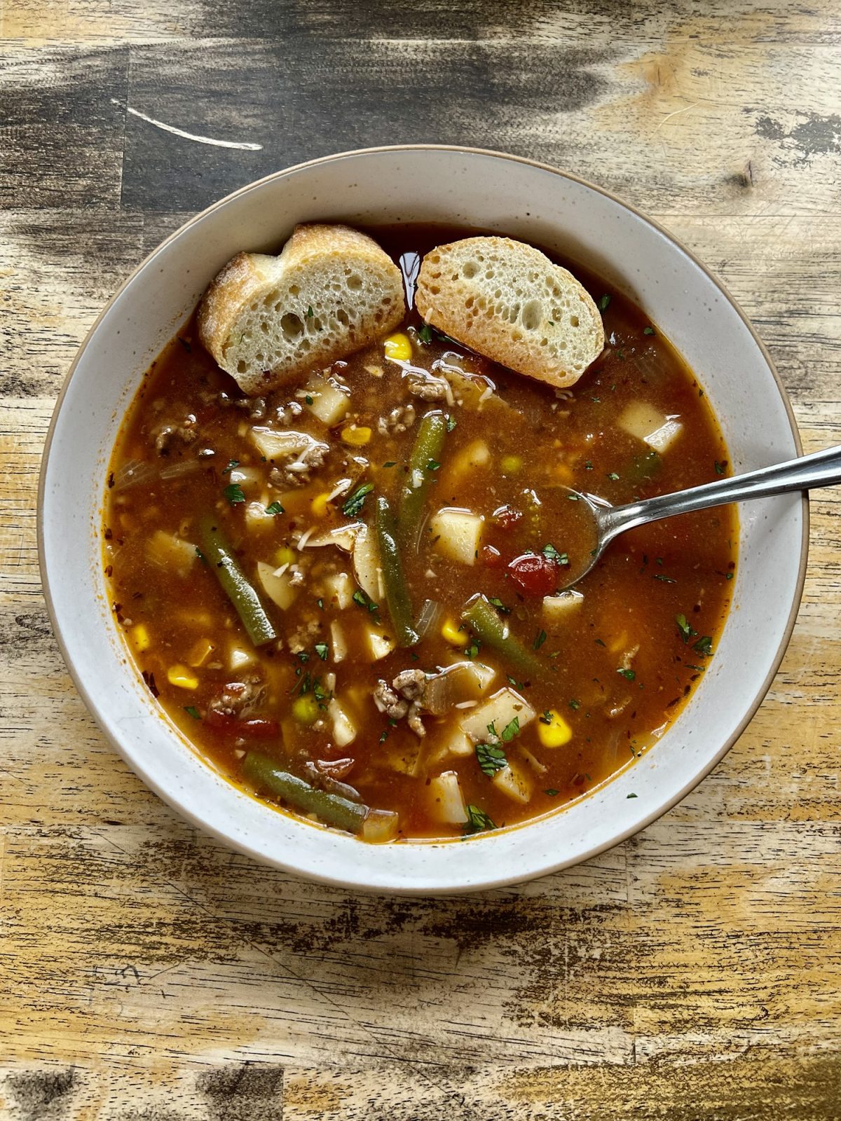 Beefy vegetable soup in a white bowl with some crispy bread and spoon inside the bowl, on a wooden table