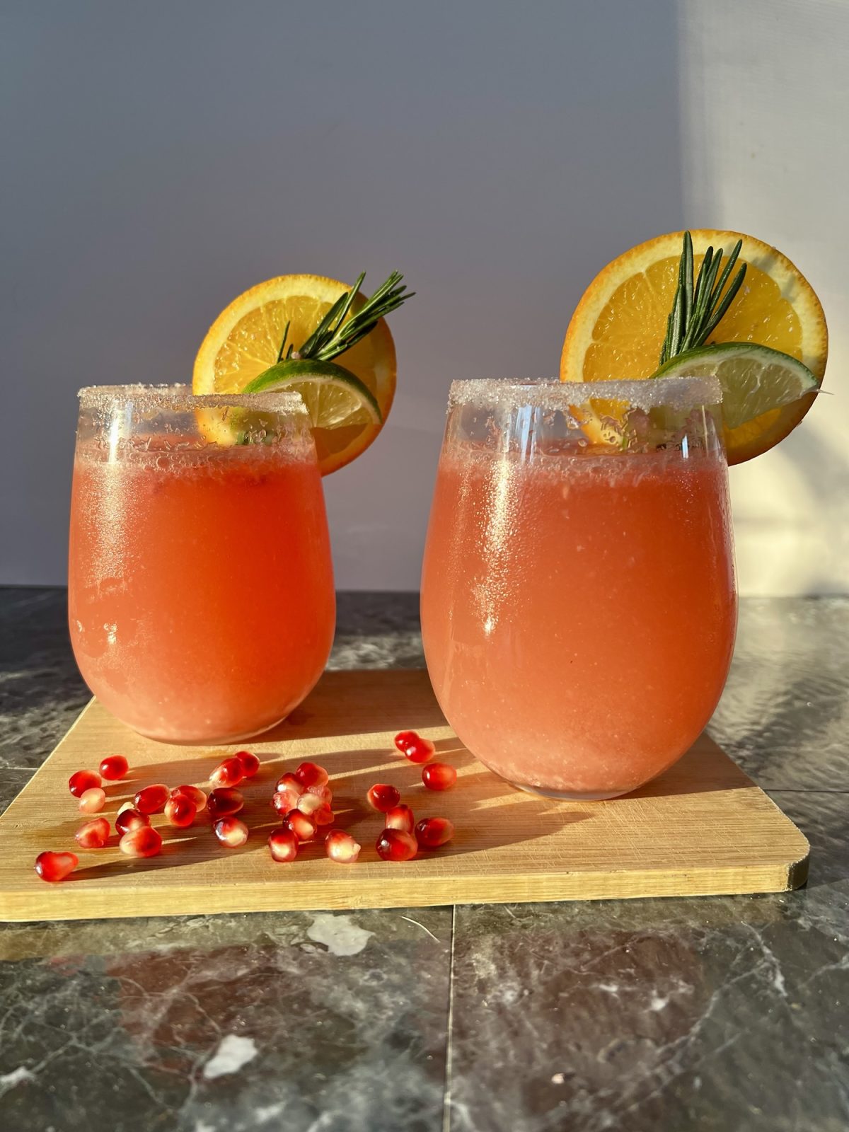Two pomegranate mimosas with fruit and rosemary for garnish on a cutting board with pomegranate seeds scattered around