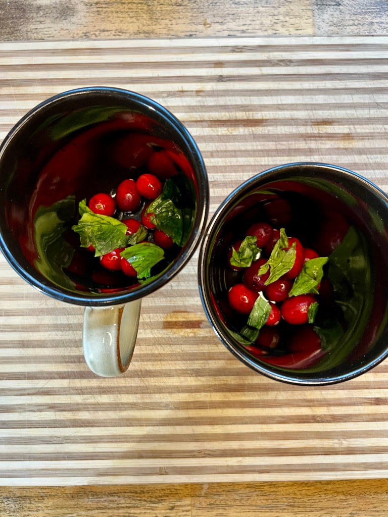Cranberries and basil in two mugs on a wooden cutting board