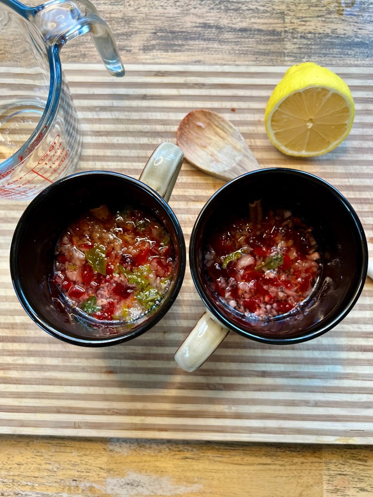 Cranberry, basil, hot water, and a cinnamon stick in some mugs with half a lemon behind