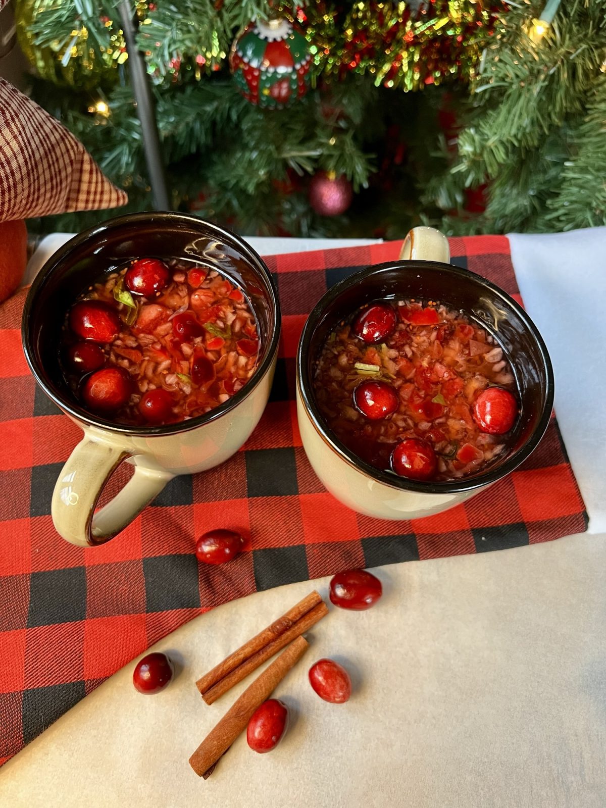 Two cranberry toddies on a table with some cinnamon sticks and cranberries scattered around with a Christmas tree behind
