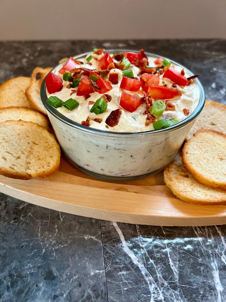 Avocado blt dip in a bowl with some bagel crisps around it on a countertop