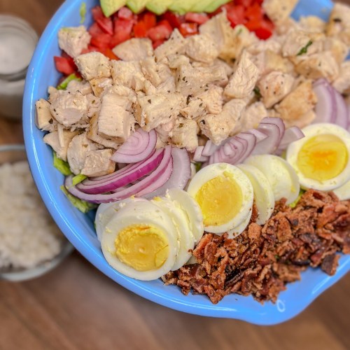 A large cobb salad in a blue bowl with blue cheese and ranch next to it in small dishes