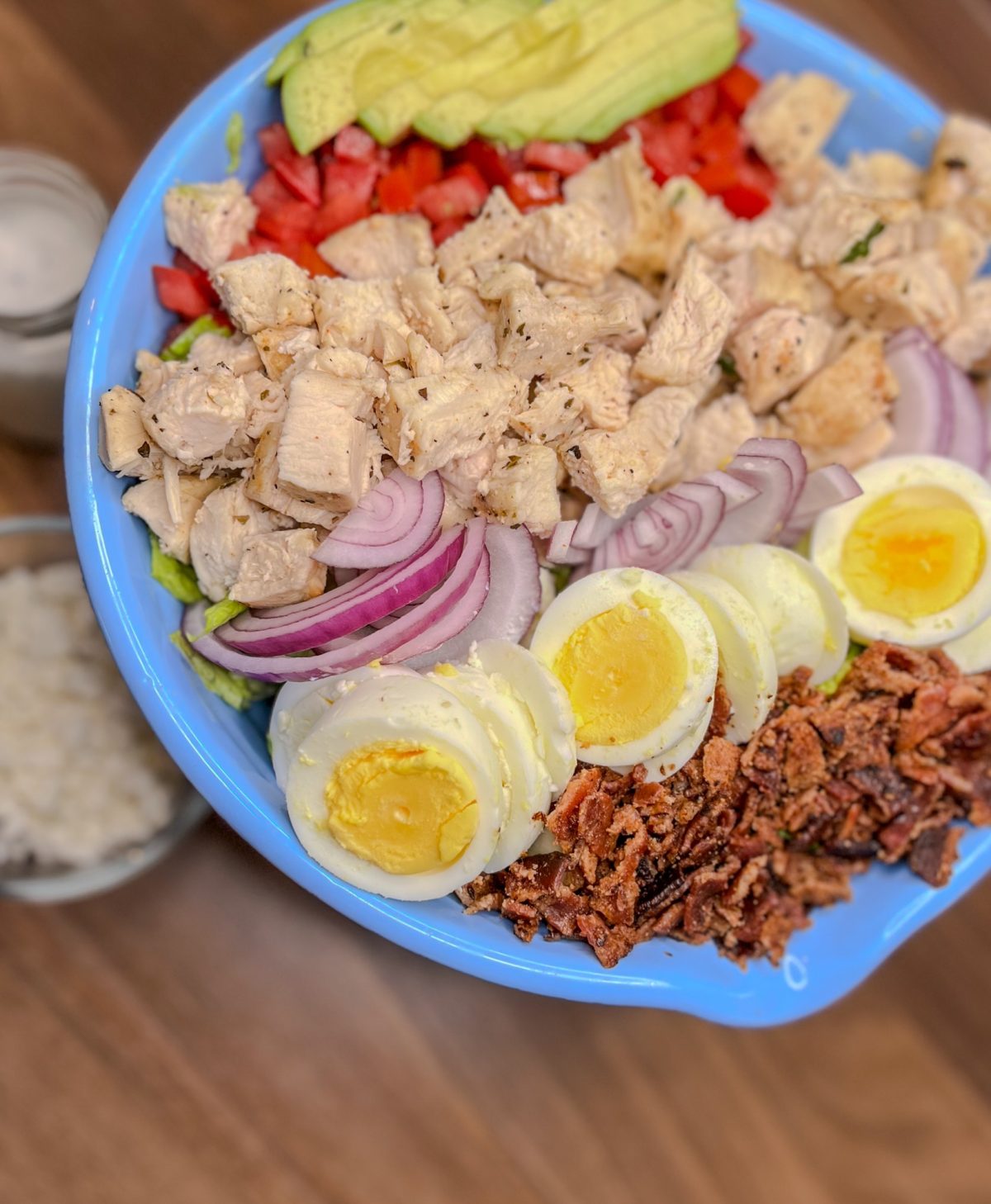 A large cobb salad in a blue bowl with blue cheese and ranch next to it in small dishes