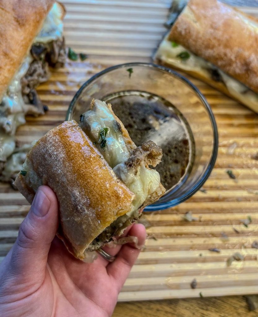 Sliced steak melt being dipped into au jus on a cutting board