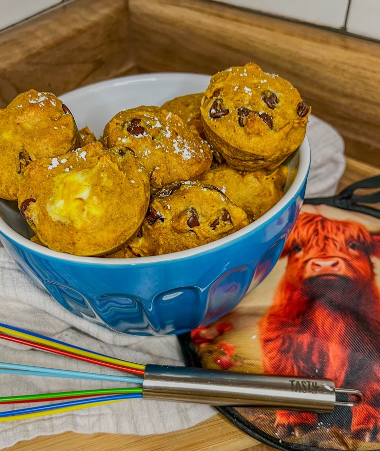 Pumpkin chocolate chip muffins in a blue bowl on a wooden countertop with a hot pad and whisk nearby