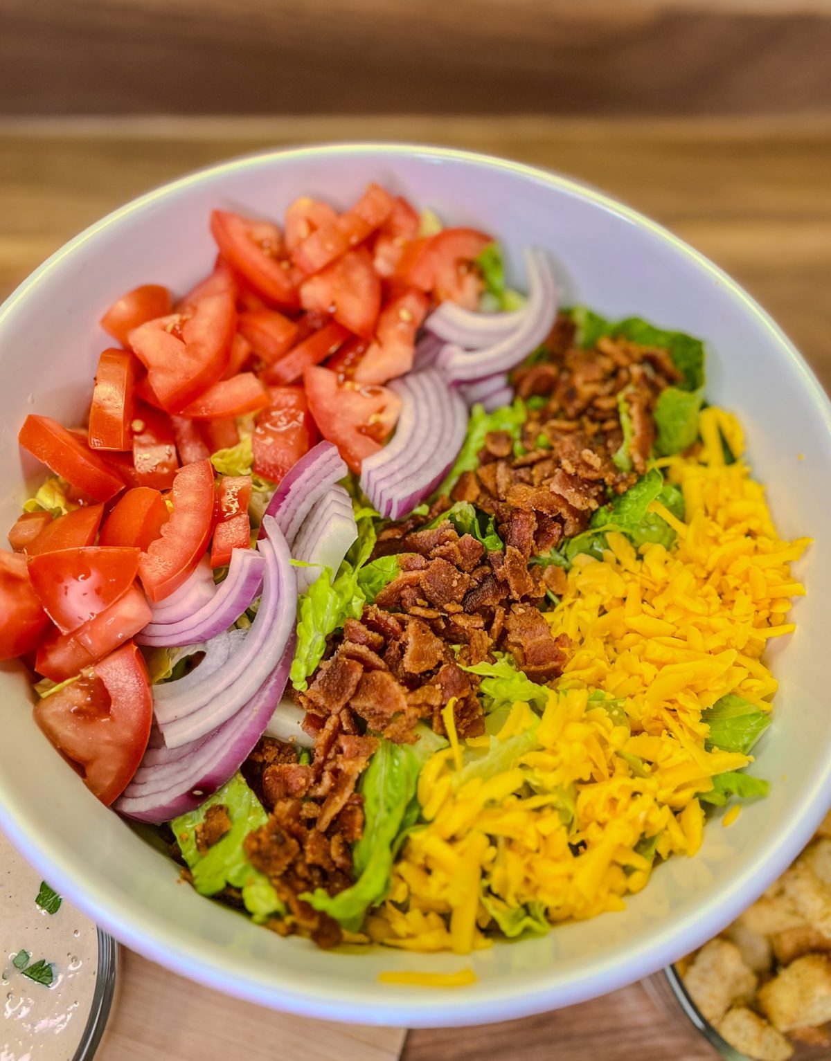 A bacon Caesar salad in a large bowl with some dressing and croutons nearby