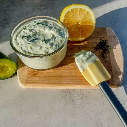 Homemade tzatziki sauce in a dish with some dill, lemon, and cucumber nearby, on a wooden cutting board