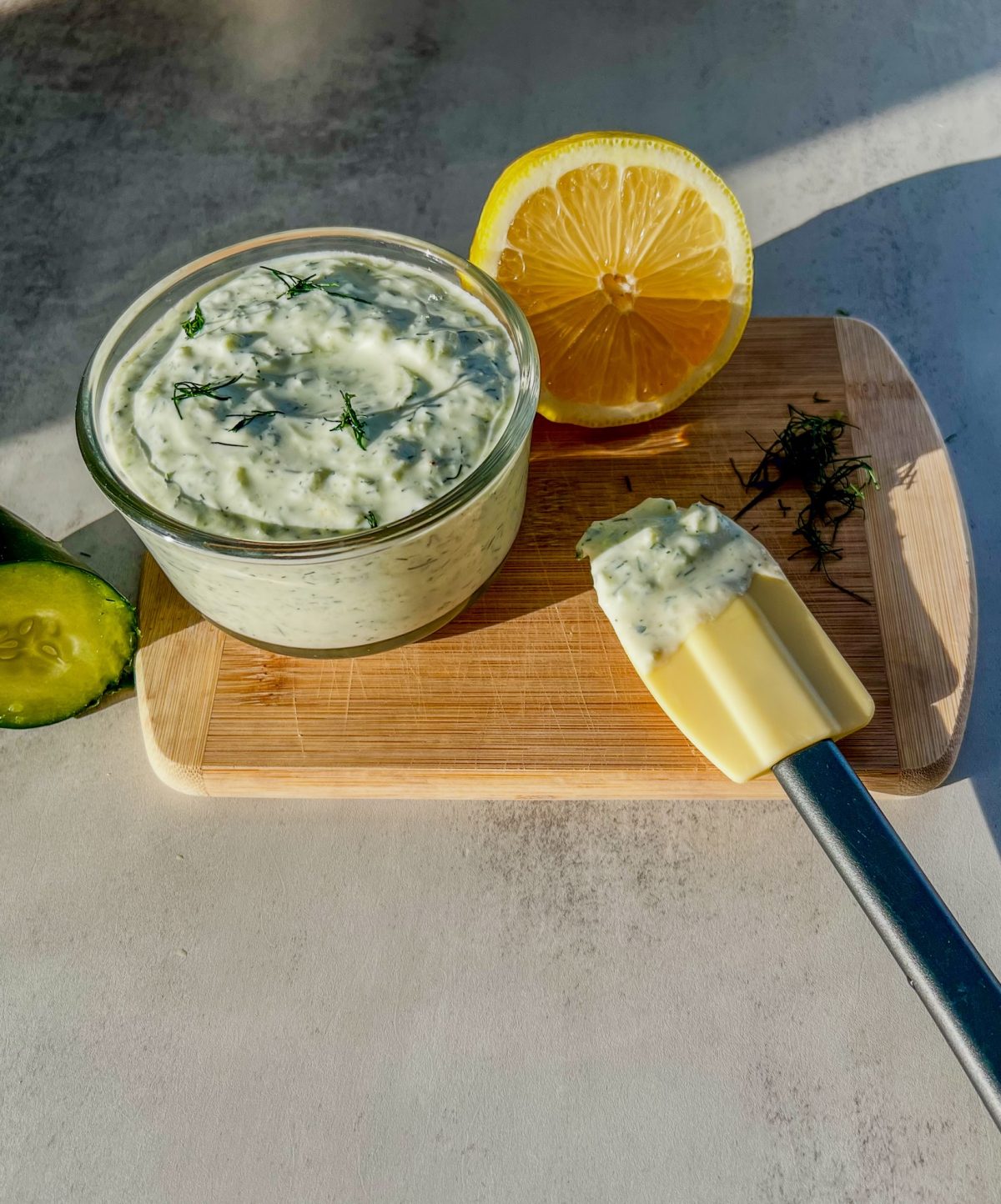 Homemade tzatziki sauce in a dish with some dill, lemon, and cucumber nearby, on a wooden cutting board