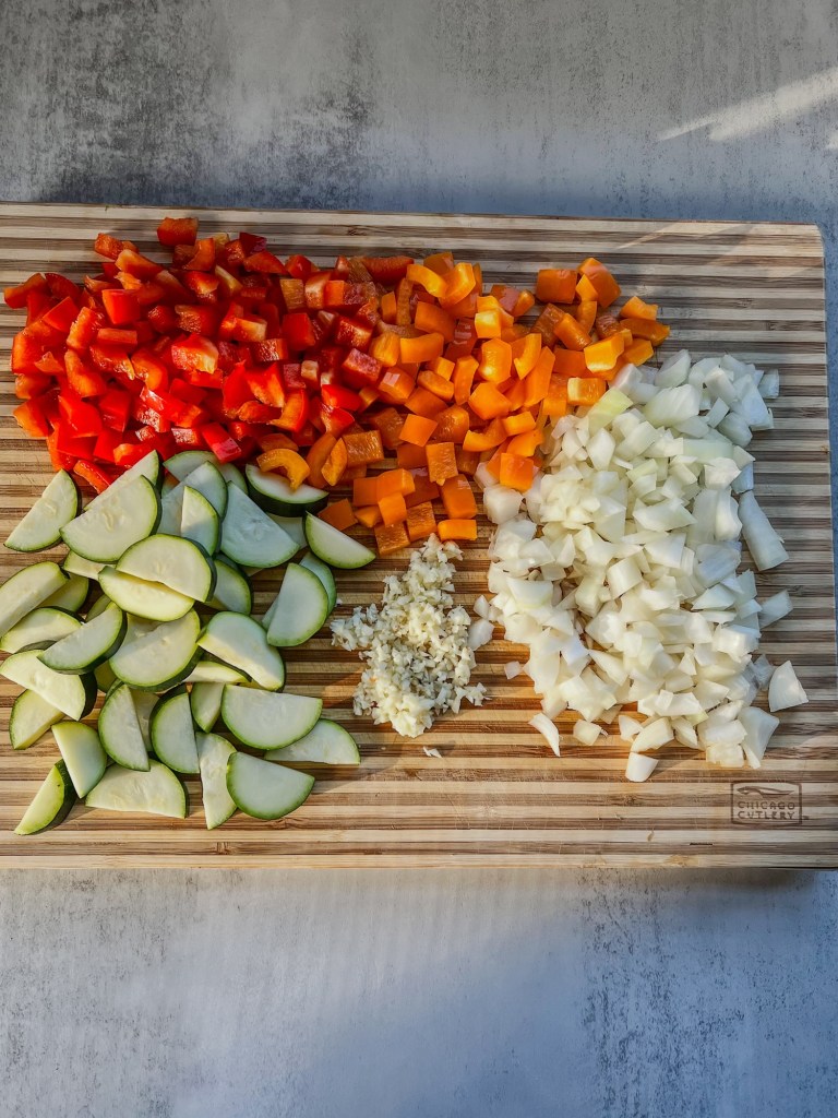 A bunch of peppers, zucchini, onion, and garlic chopped up on a cutting board