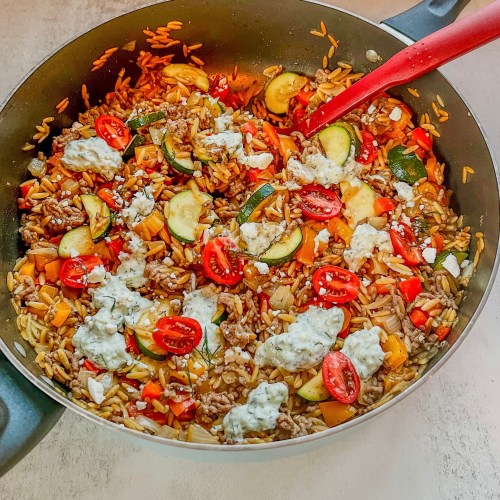 One Pot Greek Orzo in a skillet with a red serving spoon scooping some out