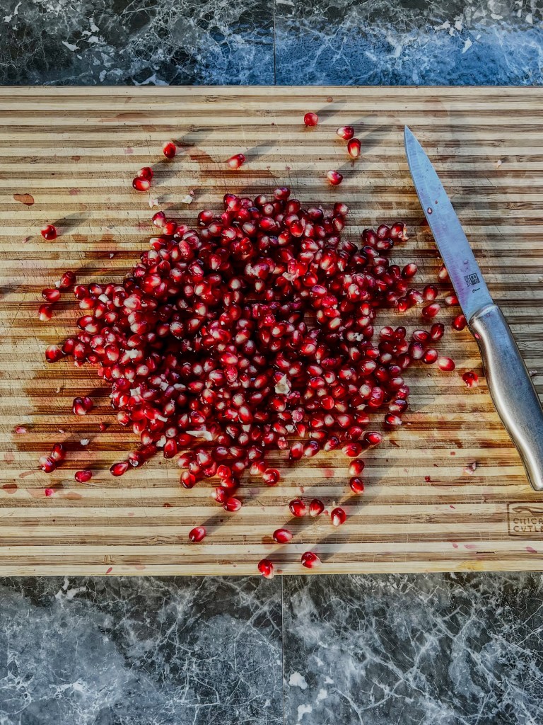 Pomegranate seeds scattered all over a wooden cutting board with a knife nearby