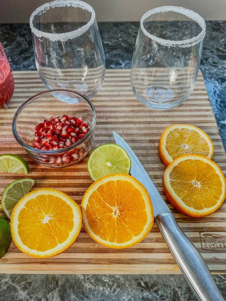 A cutting board with seeds, orange, and limes cut up, and stemless wine glasses with sugar on the rims