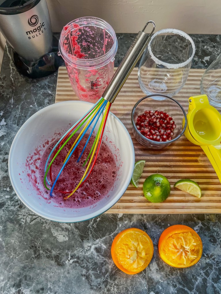 A cutting board with fruit on it and a bowl filled with fresh fruit juice with a whisk inside