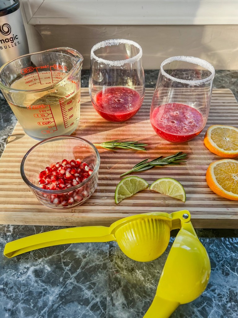 A cutting board filled with fruit, herbs, seeds, and some wine glasses