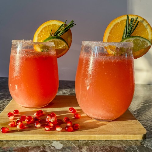 Two pomegranate mimosas with fruit and rosemary for garnish on a cutting board with pomegranate seeds scattered around