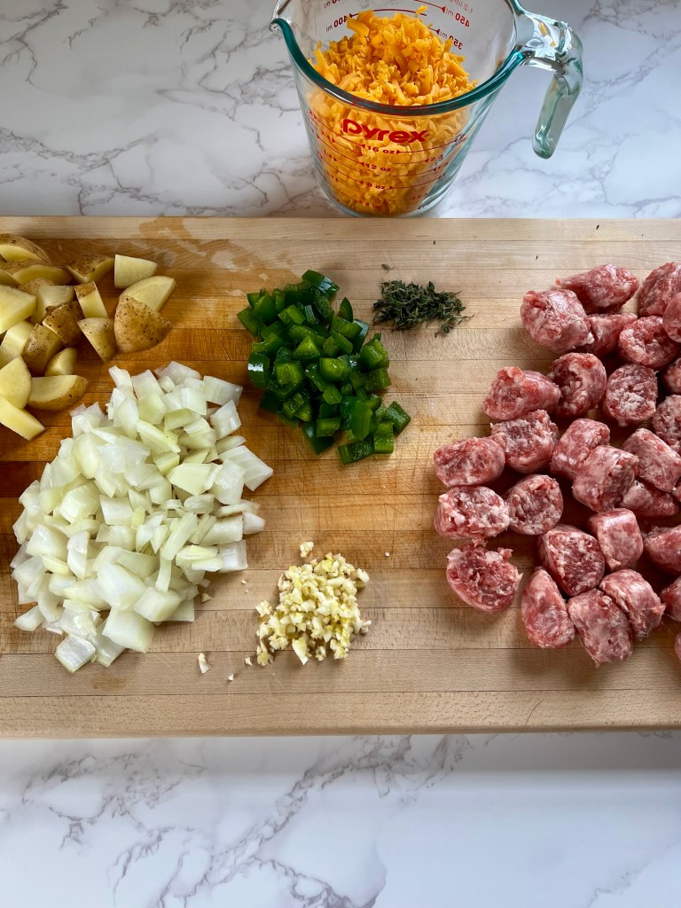 Veggies, herbs, brats, and cheese on a wooden cutting board on a countertop