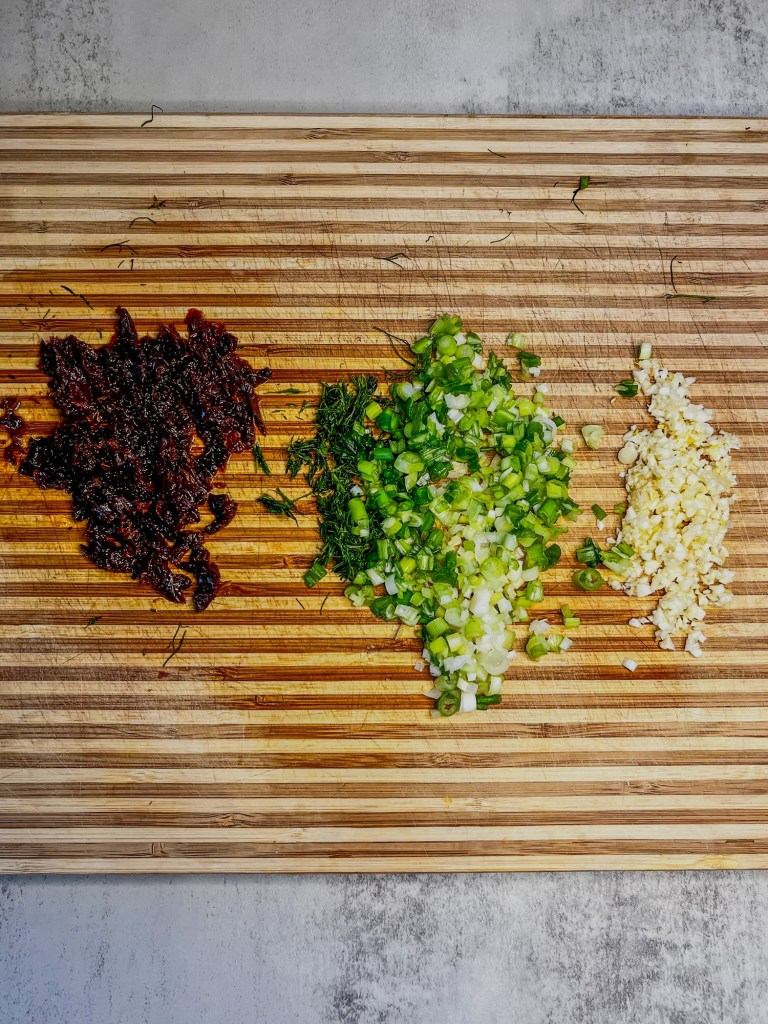 Sun-dried tomatoes, green onion, and dill, and garlic chopped up on a cutting board