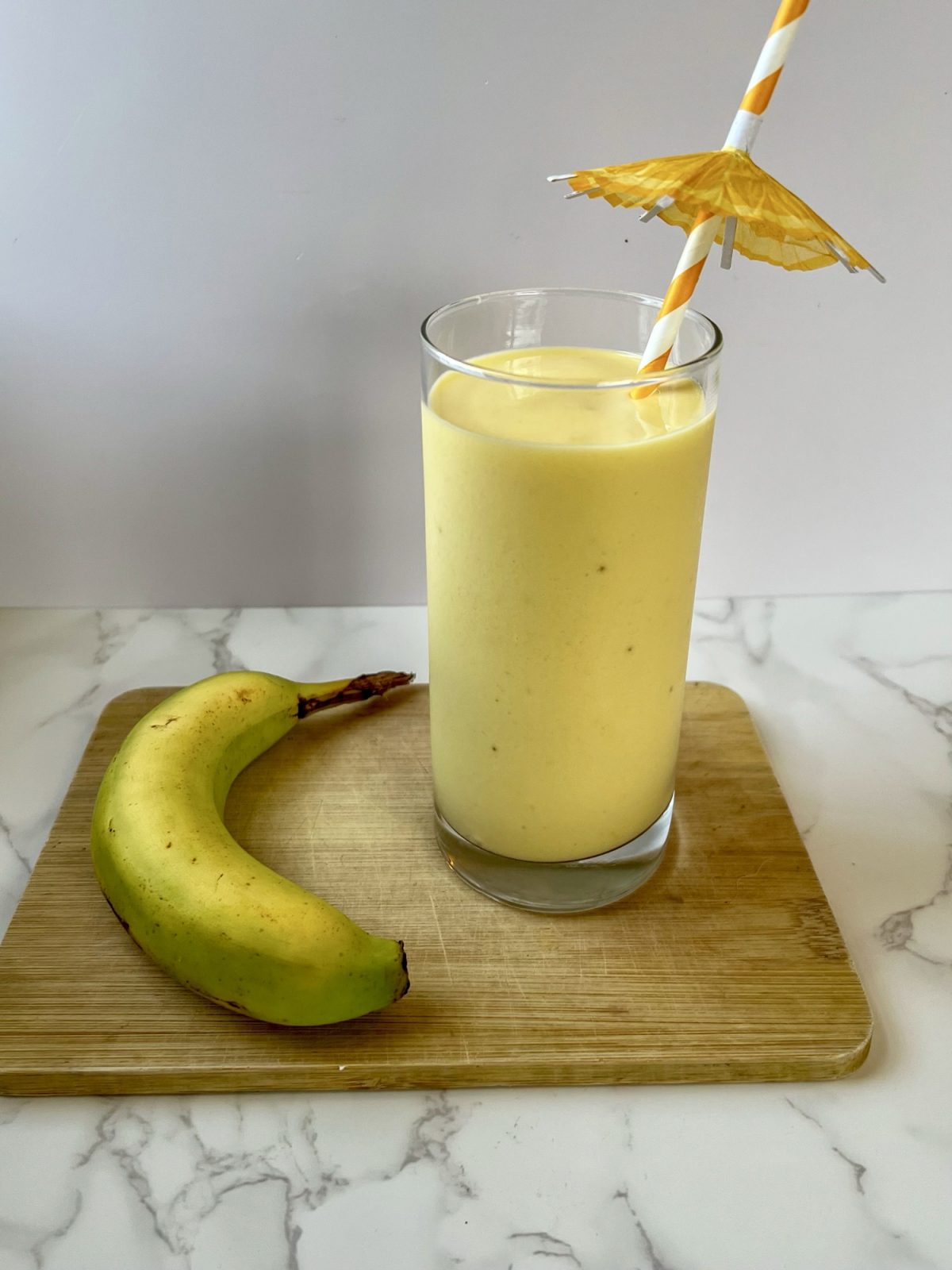 Mango Banana Smoothie in a glass with a straw in it with an umbrella, sitting on a cutting board with a banana next to it
