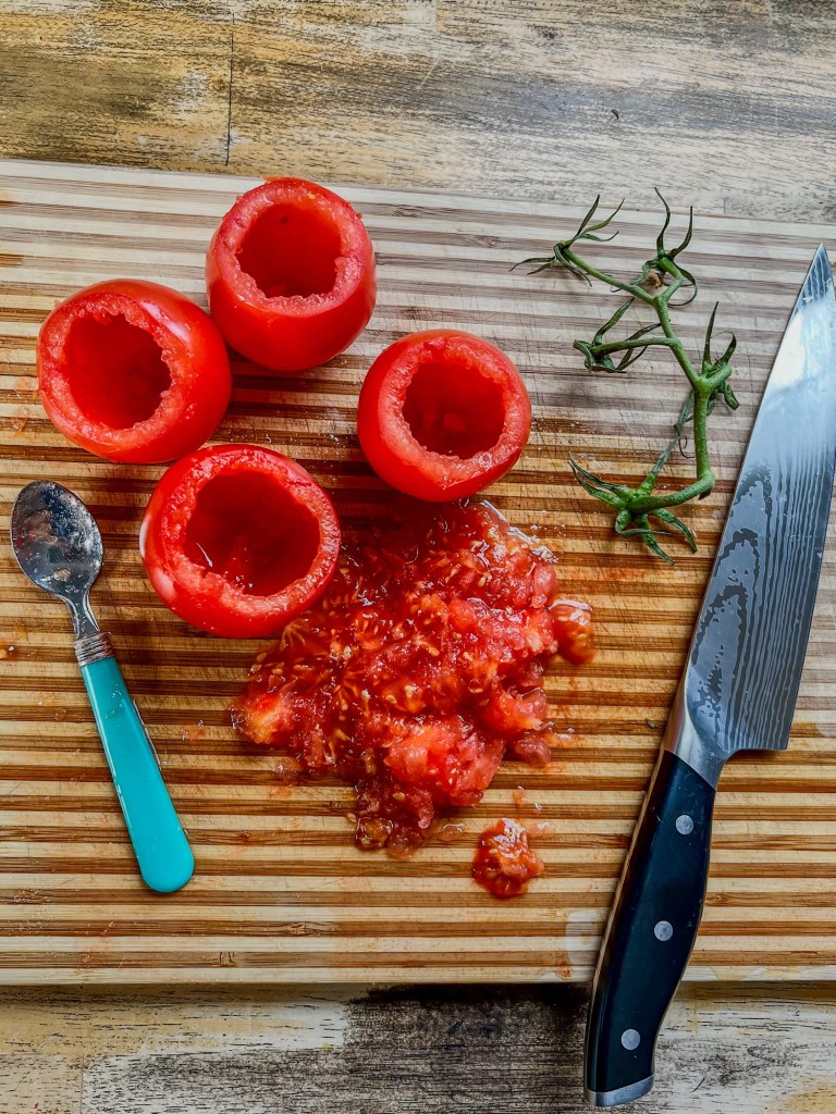 Hollowed out tomatoes on a wooden cutting board with a spoon and all the seeds nearby