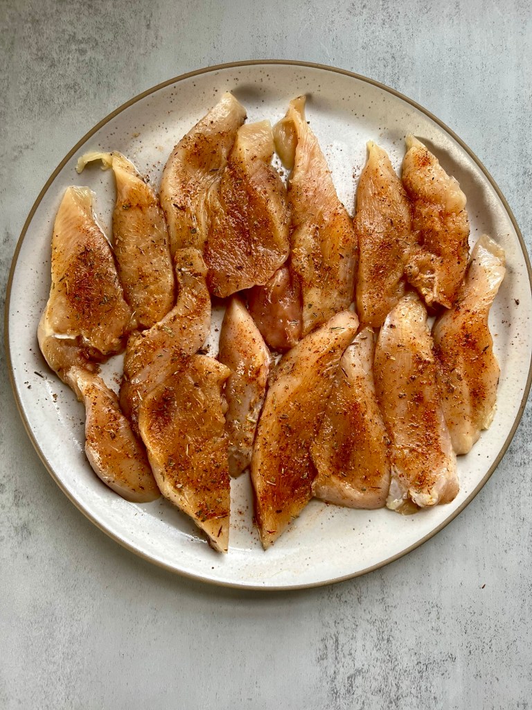 Chicken cut into tenders with a spice blend rubbed on, on a white plate