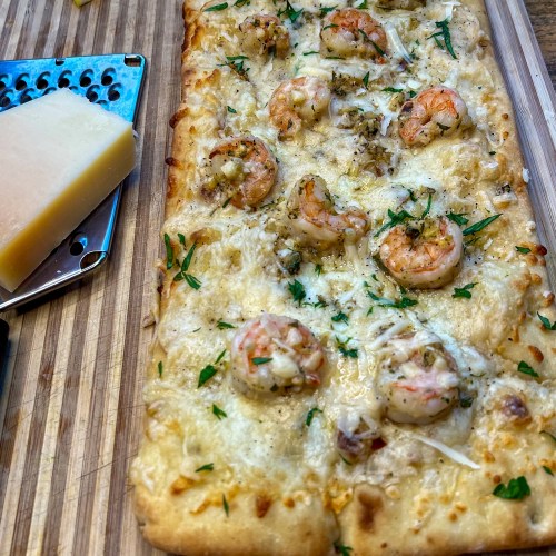Shrimp scampi flatbread on a cutting board with a cheese grater, cheese, and lemon wedges nearby