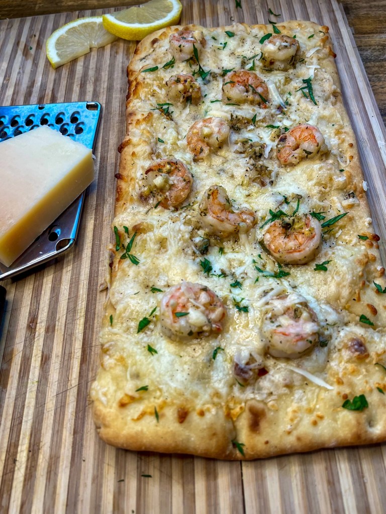 Shrimp scampi flatbread on a cutting board with a cheese grater, cheese, and lemon wedges nearby