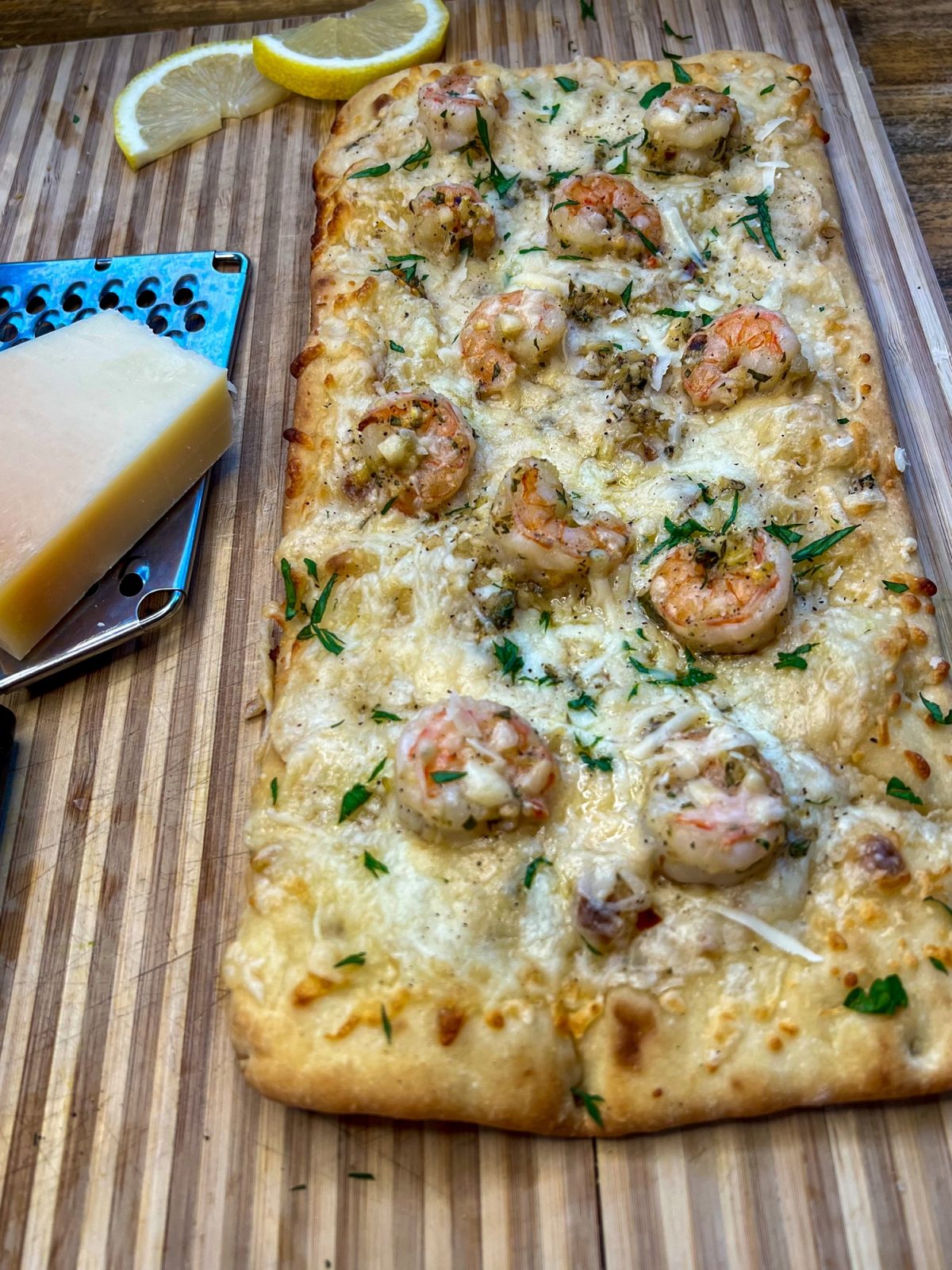 Shrimp scampi flatbread on a cutting board with a cheese grater, cheese, and lemon wedges nearby