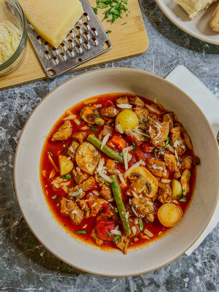 Tomato pork stew in a white bowl with some cheese and a grater nearby