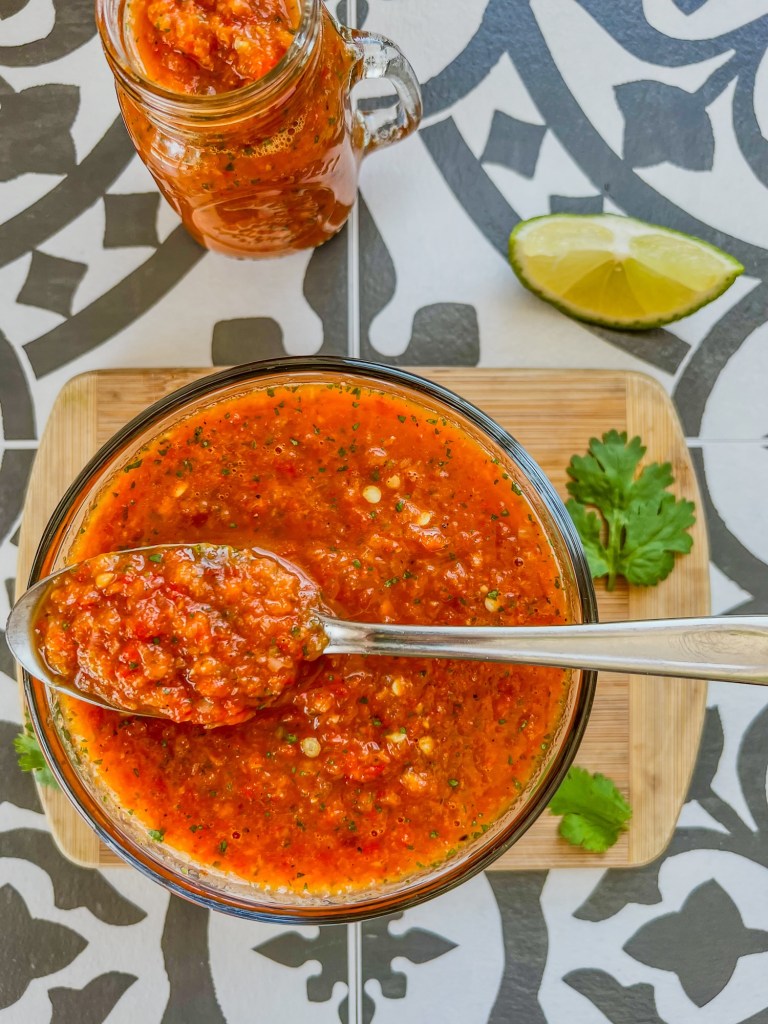 Homemade hot sauce in a dish with a spoon inside with some cilantro and lime on the side