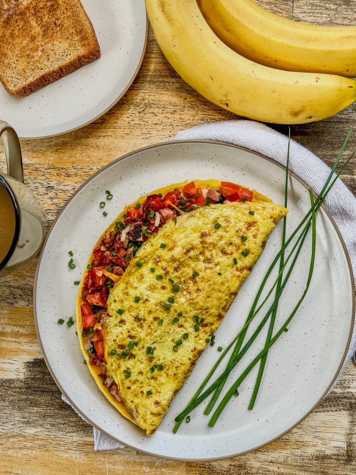 Everything omelet on a white plate with some chives for garnish, bananas, toast, and coffee around it