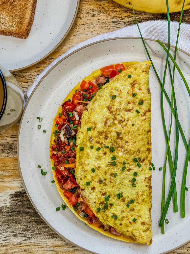 Everything omelet on a white plate with some chives for garnish, bananas, toast, and coffee around it