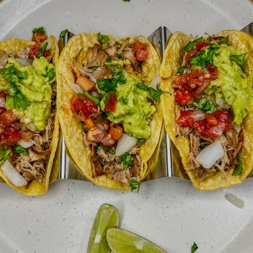 Three carnitas tacos on a white plate with some lime wedges nearby