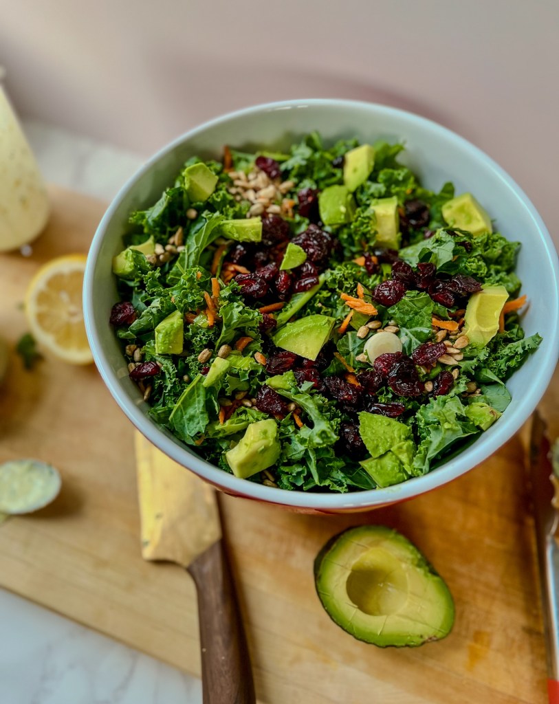 A large avocado kale salad in a bowl on a wooden cutting board with veggies and a knife nearby