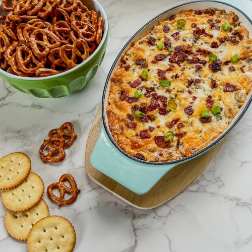 Bacon Jalapeño Popper dip in a glass dish with a bowl of pretzels behind it and some crackers laying around