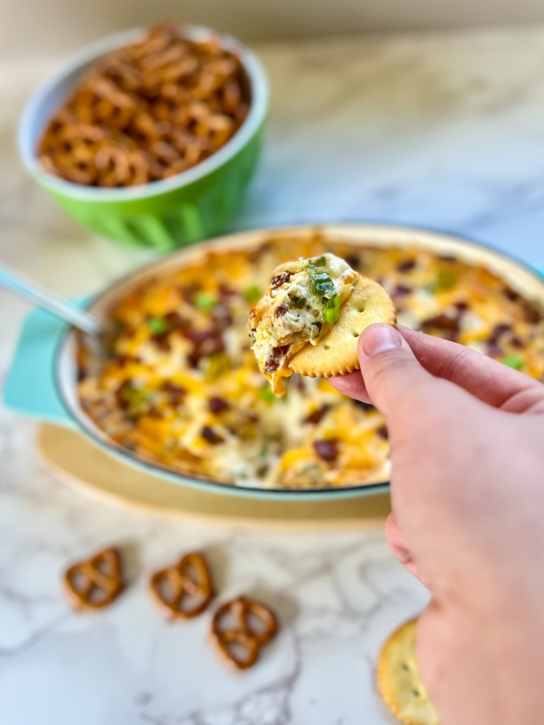 Bacon jalapeño popper dip in a dish with some pretzels and crackers nearby with some being scooped out and held up