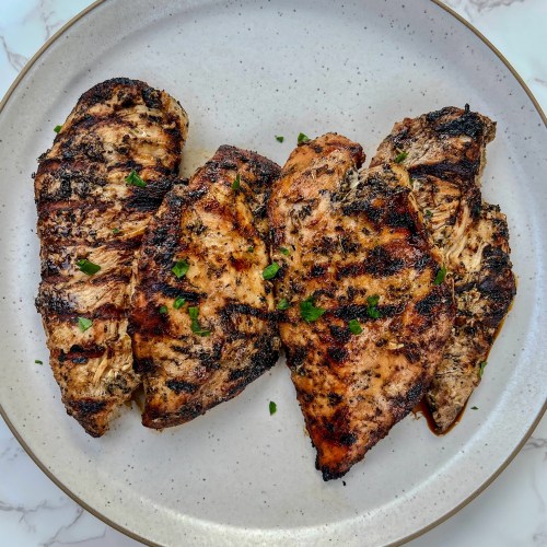 Four chicken breasts that have been marinated and grilled on a white plate, on a marble countertop