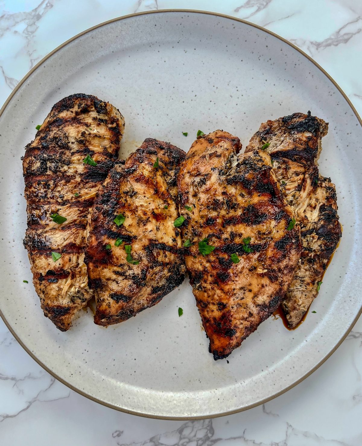 Four chicken breasts that have been marinated and grilled on a white plate, on a marble countertop