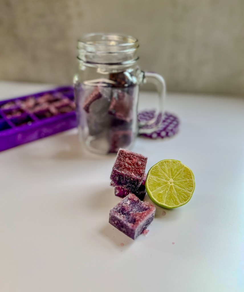 Boozy blackberry ice cubes stacked up with some lime next to it and a glass full of the cubes and an ice cube tray behind