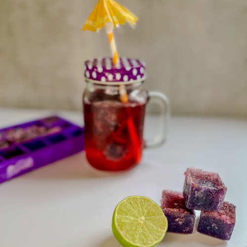 Boozy blackberry cubes stacked up with some lime next to it and a drink and ice cube tray behind