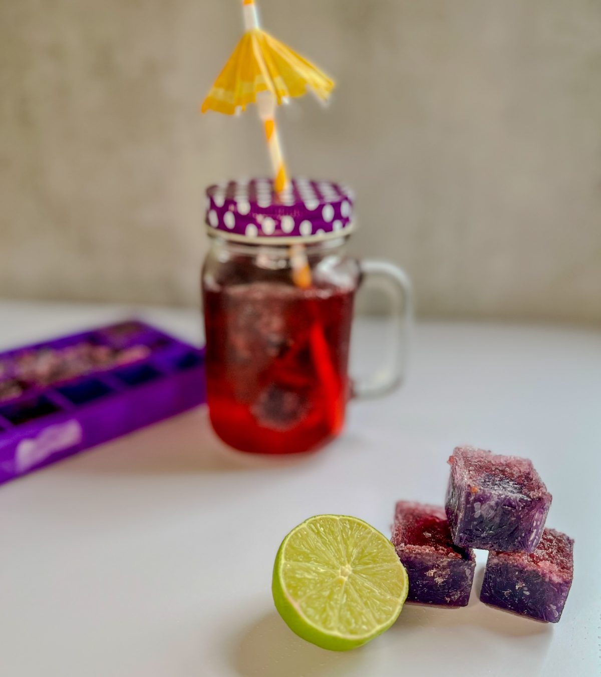 Boozy blackberry cubes stacked up with some lime next to it and a drink and ice cube tray behind